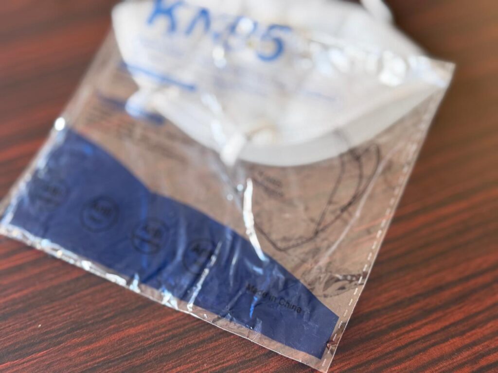 A factory-sealed white KN95 protective face mask in its clear plastic retail packaging on a dark wood surface