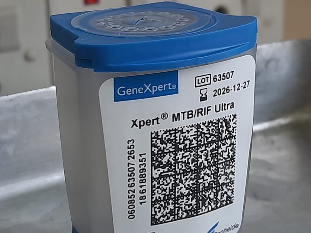 The image shows a single GeneXpert® Xpert® MTB/RIF Ultra diagnostic cartridge. It features a blue flip-top lid, a clear plastic body, and a white label with a QR code. The label indicates a LOT number of 63507 and an expiry date of 2026-12-27.